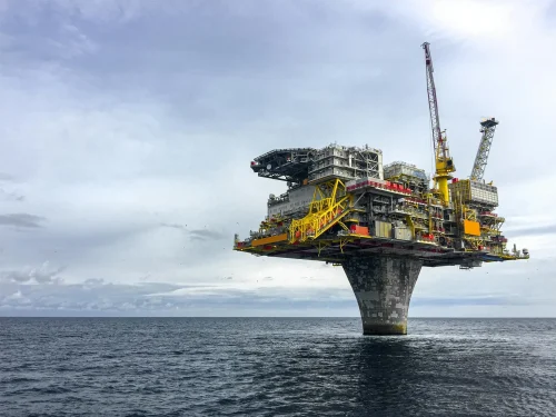 An offshore oil platform elevated above the water, supported by a single concrete column, with various industrial structures and cranes on top, set against a cloudy sky.